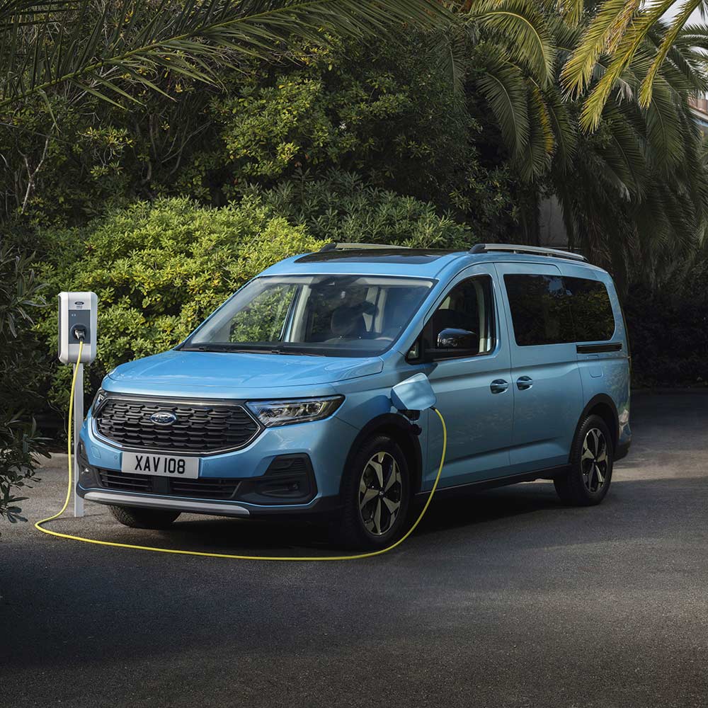 Světle modrý Ford Tourneo Connect se nabíjí na příjezdové cestě, obklopený zelenou vegetací a palmami.
