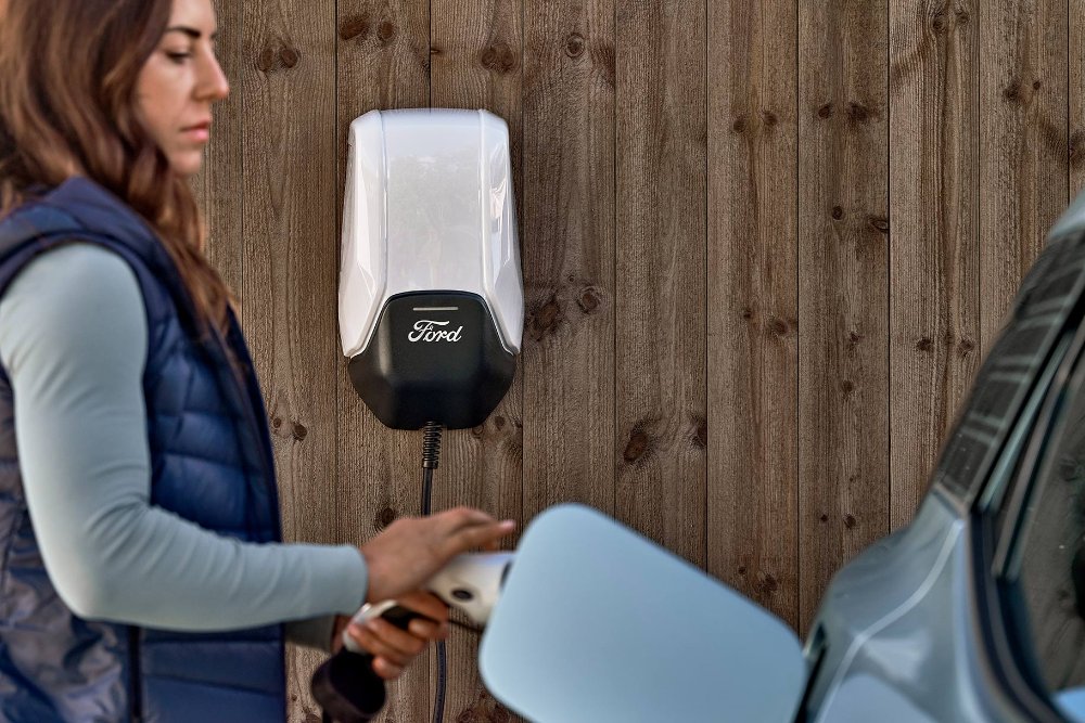 Žena připojuje své modré elektrické vozidlo Ford Explorer k domácí nabíjecí stanici.