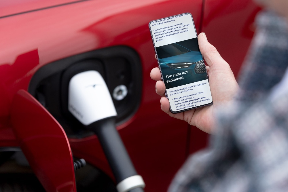 Člověk stojí vedle elektrického vozidla u nabíjecí stanice a čte si na mobilním telefonu článek vysvětlující Akt EU o datech.