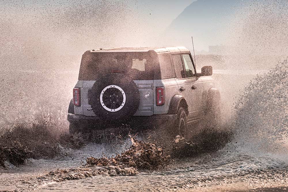 Pohled zezadu na stříbrný Ford Bronco, který projíždí vodní tříští na pláži.