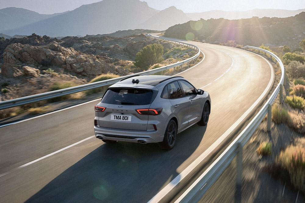 Stříbrný Ford Kuga projíždí malebnou trasou, v pozadí slunečná horská krajina.