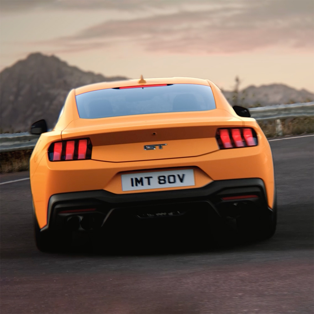 Zadní část Ford Mustang GT Fastback v barvě Orange Fury projíždějící ostrou zatáčku na venkovské silnici.