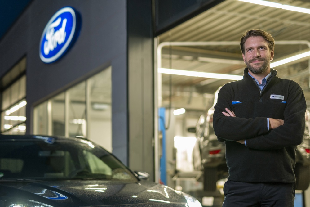 Usmívající se technik Ford se zkříženýma rukama stojí v servisním středisku. Na budově je logo Ford a v popředí je auto.