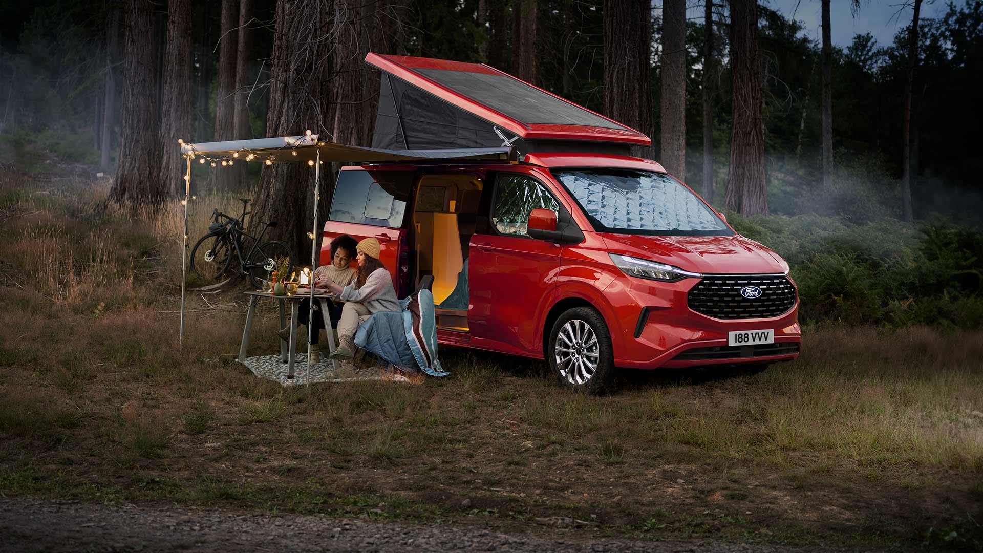 Two women sit under an awning from the open side doors of a red Ford Transit Custom Nugget, with the tilt roof up.