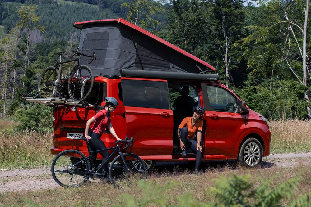 Lidé sundavají kola z nosiče upevněného na červeném obytném voze Ford Transit Custom Nugget.