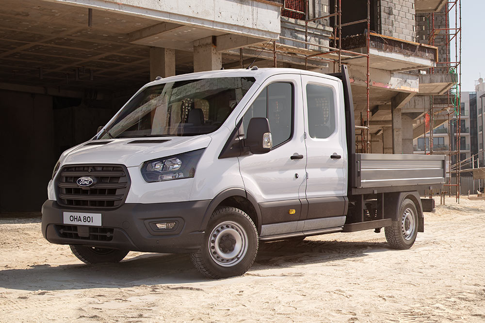 Bílý Ford Transit Custom s podvozkem s kabinou a valníkem je zaparkovaný na staveništi.