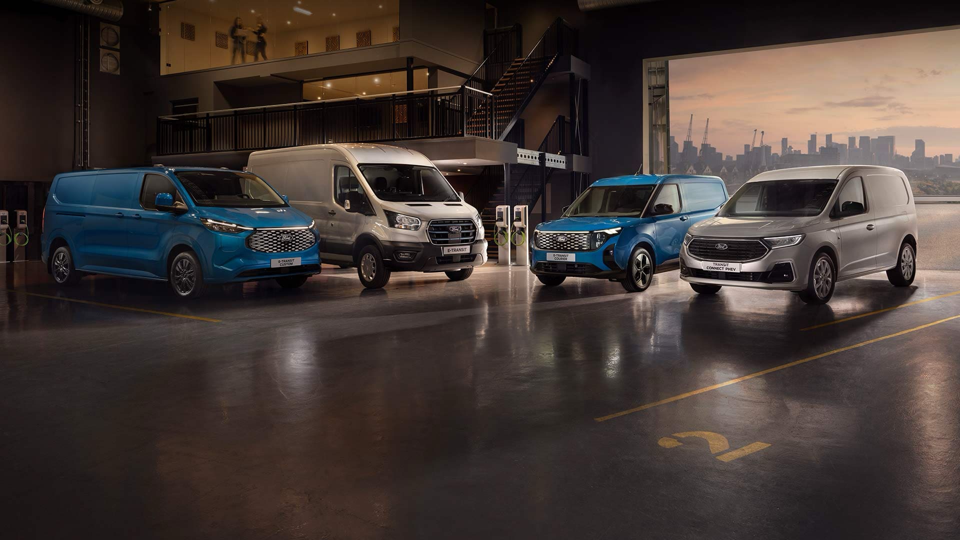 Flotila vozidel Ford Transit ve showroomu s panoramatem města za soumraku v pozadí.