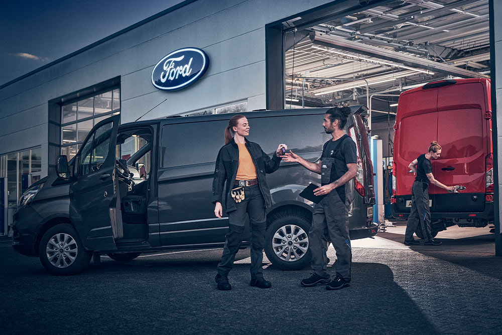 Dva technici si předávají klíče před servisní dílnou Ford s dodávkami na pozadí.
